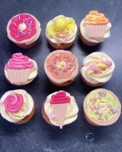 Neuf cupcakes décorés de façon ludique avec des glaçages représentant des donuts, cornets de glace et tourbillons roses, jaunes et blancs sur fond noir.