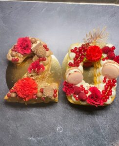 Gâteau '20' en forme de chiffres, décoré de crème, fleurs rouges, macarons, fruits rouges (groseilles, framboises) et Ferrero Rocher sur fond sombre.