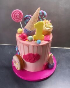 Gâteau d'anniversaire rose pour un an, décoré de bonbons, sucettes, cône de glace, et un grand chiffre '1' jaune.