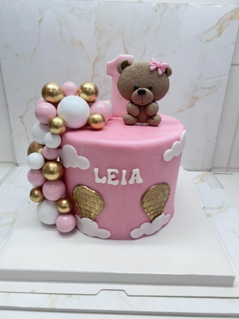 Gâteau d'anniversaire rose pour la première année de Leia, décoré d'un ourson, d'un grand '1', de balles roses/blanches/dorées et de montgolfières dorées.