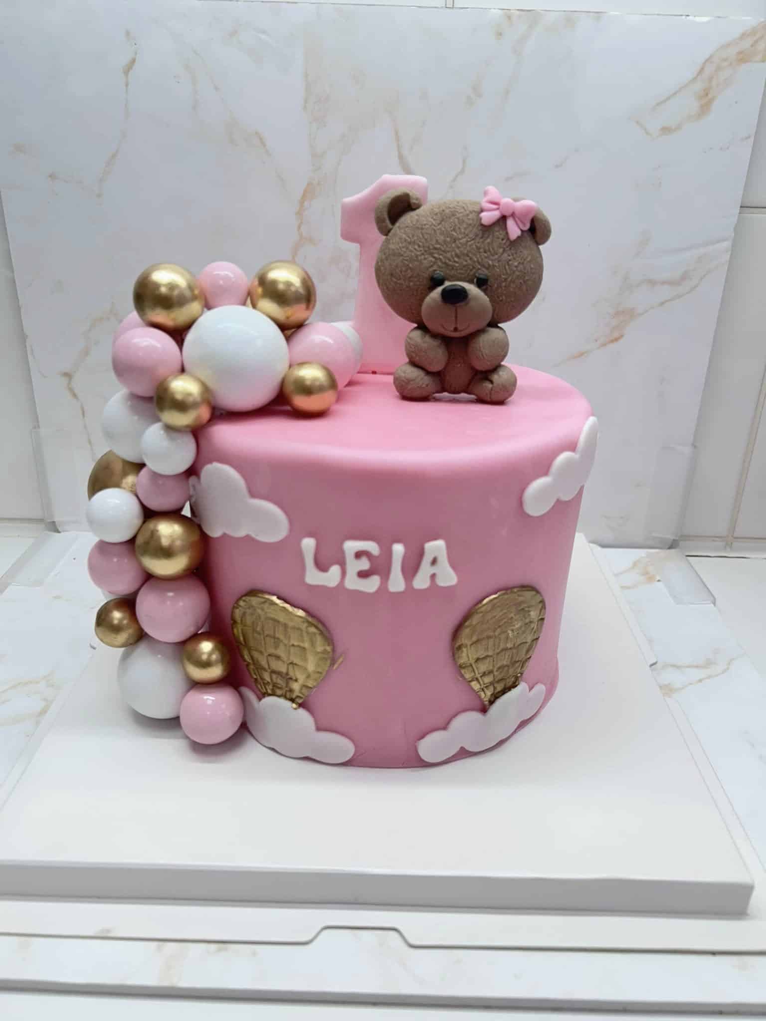 Gâteau d'anniversaire rose pour la première année de Leia, décoré d'un ourson, d'un grand '1', de balles roses/blanches/dorées et de montgolfières dorées.