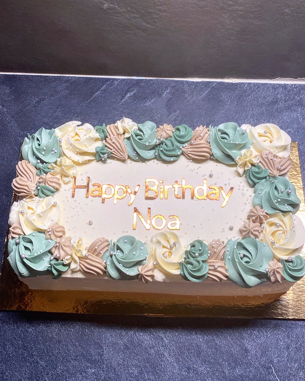 Gâteau d'anniversaire rectangulaire avec glaçage blanc, décoré de rosettes vert sauge, crème et beige, portant l'inscription dorée 'Happy Birthday Noa'.