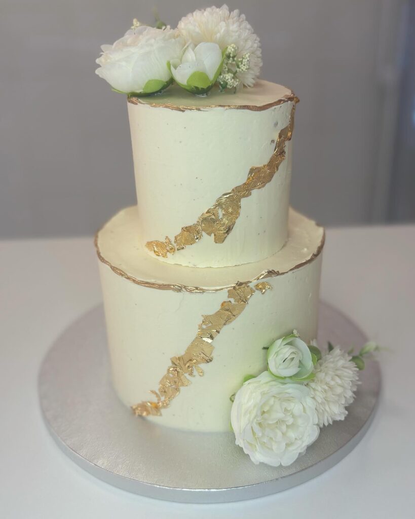 Gâteau deux étages crème et or : glaçage blanc cassé texturé avec feuille d'or en diagonale et fleurs blanches artificielles.
