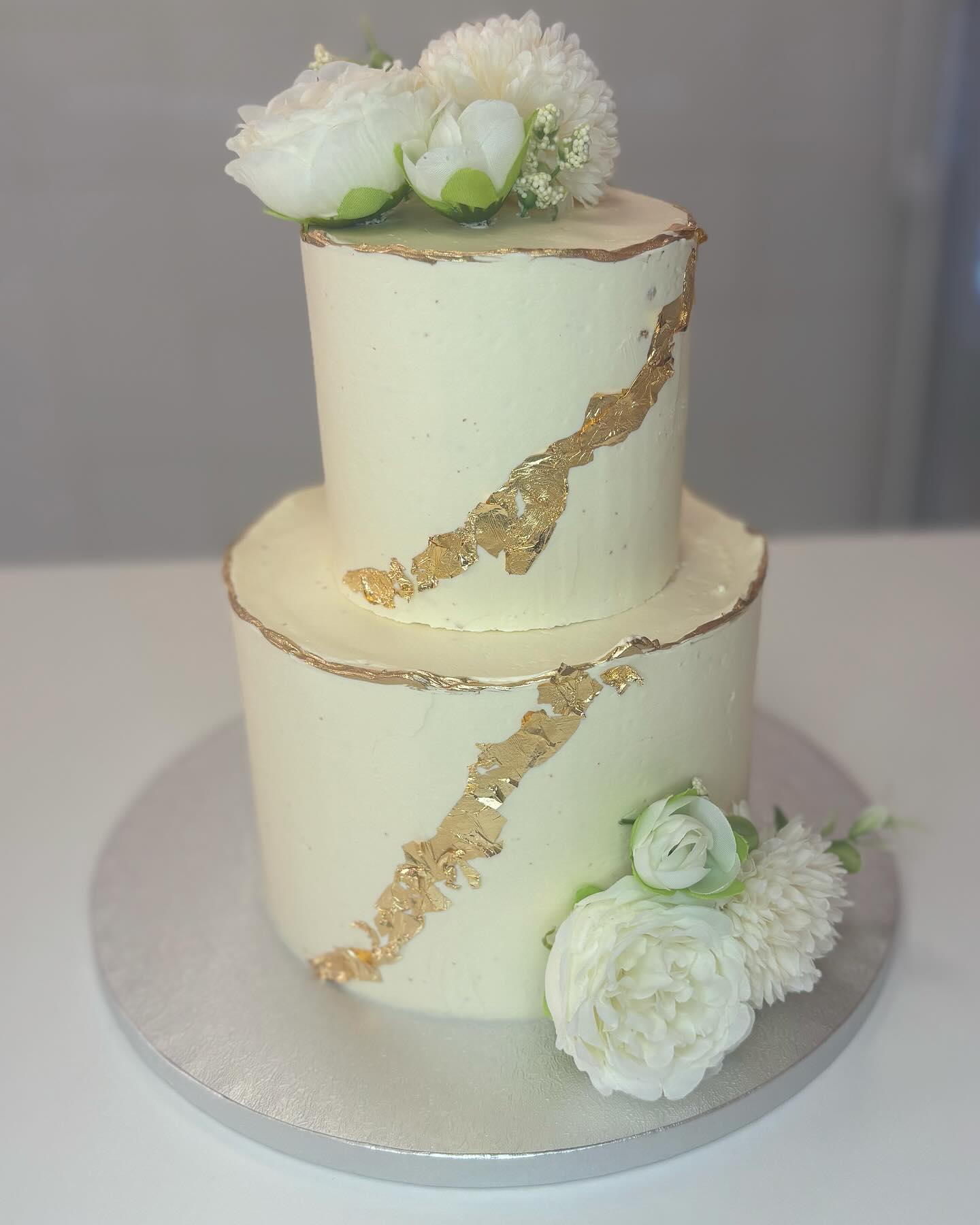 Gâteau deux étages crème et or : glaçage blanc cassé texturé avec feuille d'or en diagonale et fleurs blanches artificielles.
