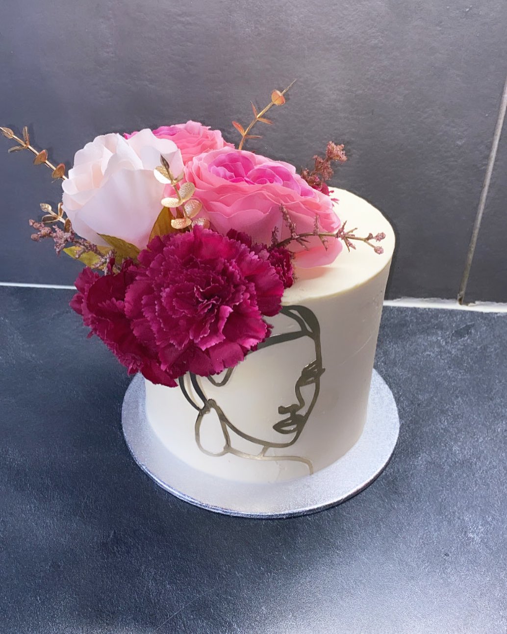 Gâteau blanc décoré d'un dessin au trait doré d'un visage de femme, surmonté d'un arrangement floral de roses roses pâles et fuchsia et d'un œillet bordeaux.
