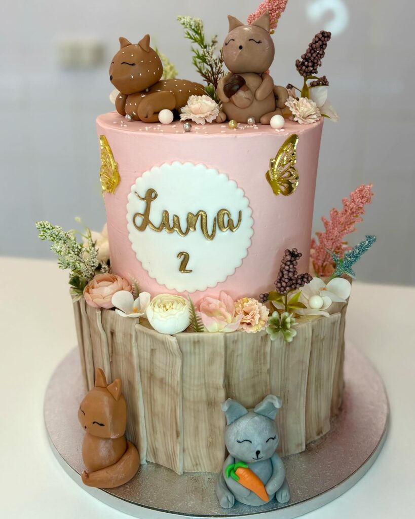 Gâteau d'anniversaire deux étages rose et tronc d'arbre, décoré de figurines d'animaux de la forêt, d'une étiquette "Luna 2" et de fleurs.