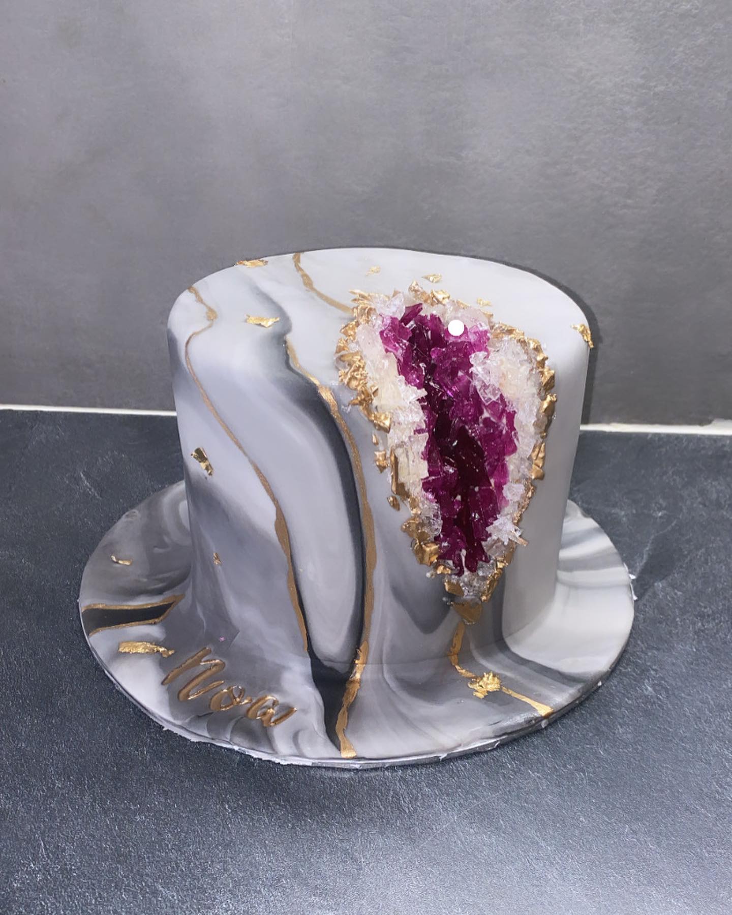 Gâteau cylindrique effet marbre gris et blanc avec veines dorées, orné d'une géode en sucre violet vif et cristaux blancs, signature 'Nora' dorée sur le socle.