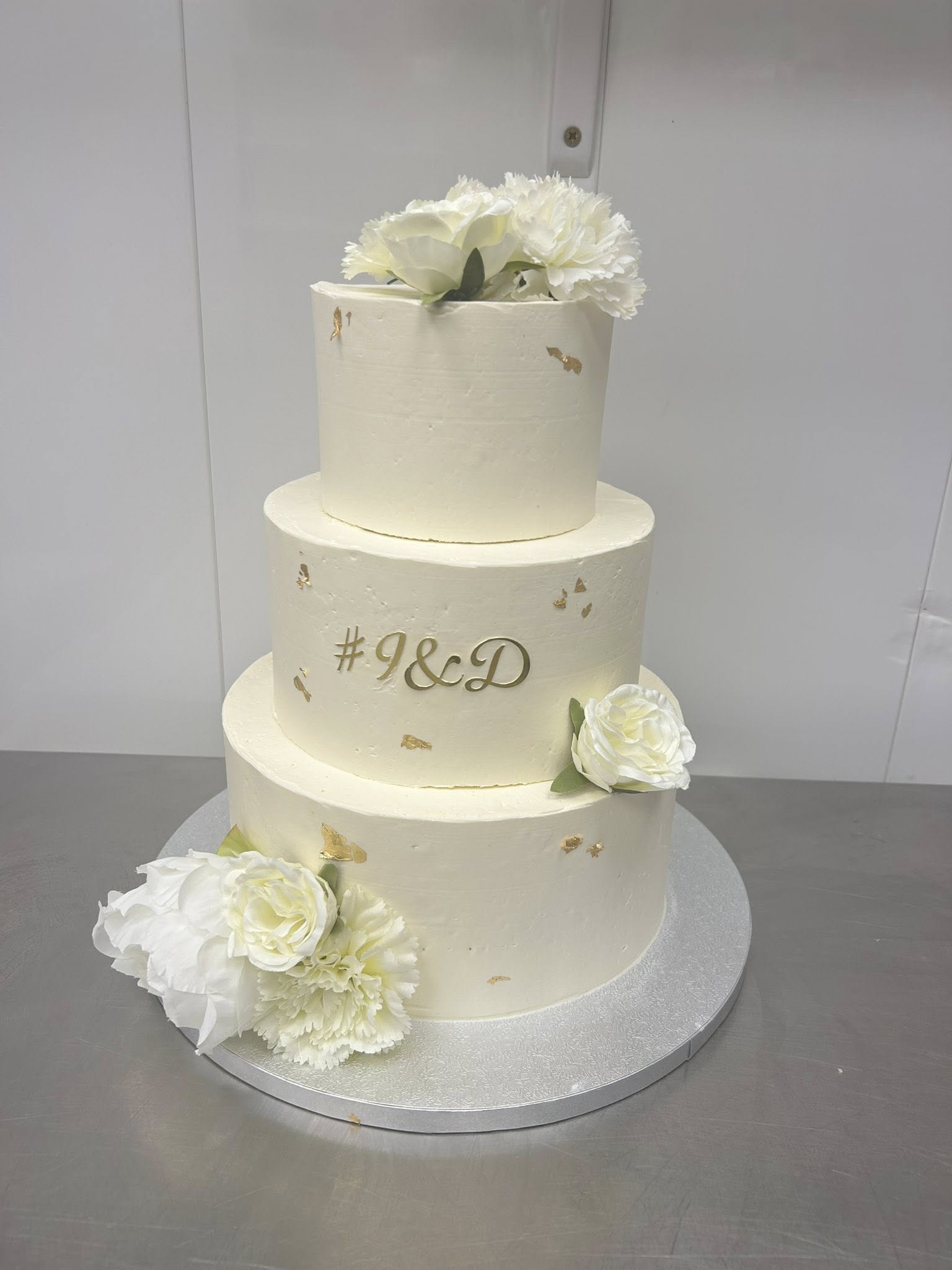 Gâteau de mariage 3 étages blanc cassé orné de fleurs, feuilles d'or et inscription dorée #J&D.
