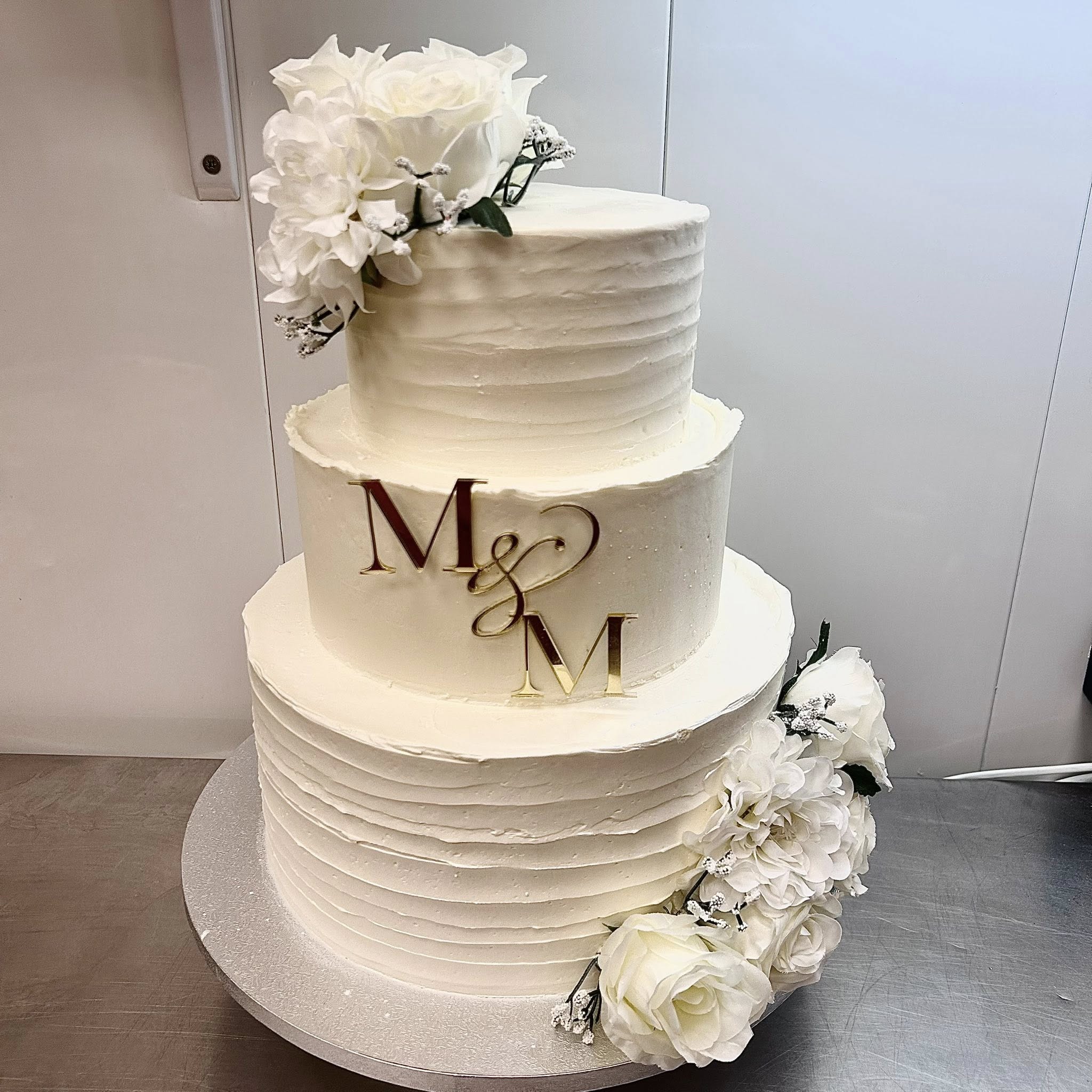 Gâteau de mariage à trois étages, fleurs blanches, glaçage texturé et monogramme doré M & M élégant.