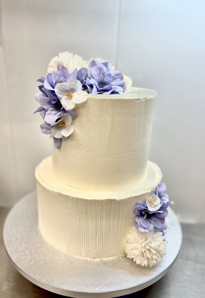 Gâteau de Mariage à Deux Étages avec Fleurs Violettes Gâteau à deux étages blanc avec glaçage texturé et fleurs artificielles violettes et blanches.