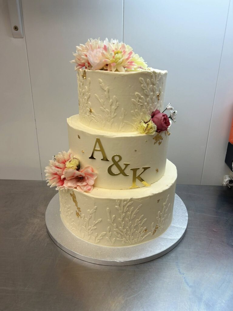 Gâteau de Mariage 3 Étages Blanc et Or avec Initiales A&K Gâteau de mariage à trois étages blanc, décorations florales, feuilles d'or et initiales A&K dorées.