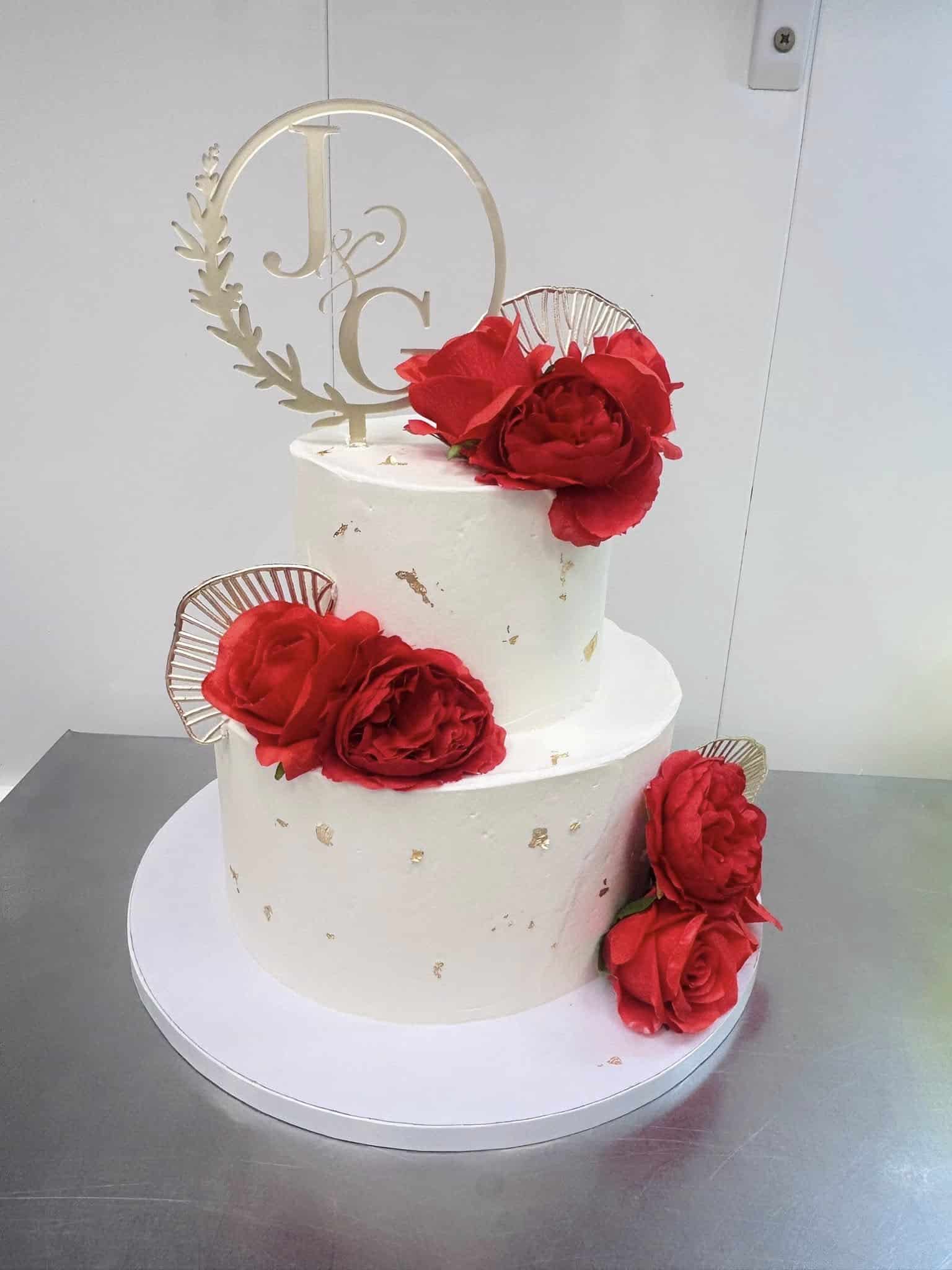 Gâteau de mariage blanc à deux étages avec roses rouges, feuilles d'or et topper doré "J&G" élégant.