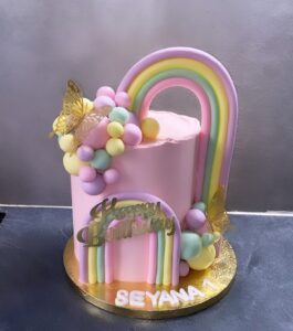 Gâteau d'anniversaire rose décoré d'un grand arc-en-ciel pastel, de balles multicolores, de papillons dorés et du texte "Happy Birthday Seyana 1".