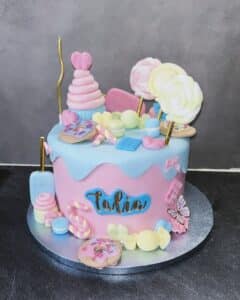 Gâteau d'anniversaire rose et bleu ciel, style "dripping" et surchargé de décorations de bonbons, macarons, beignets et sucettes. Inscription "Talia".