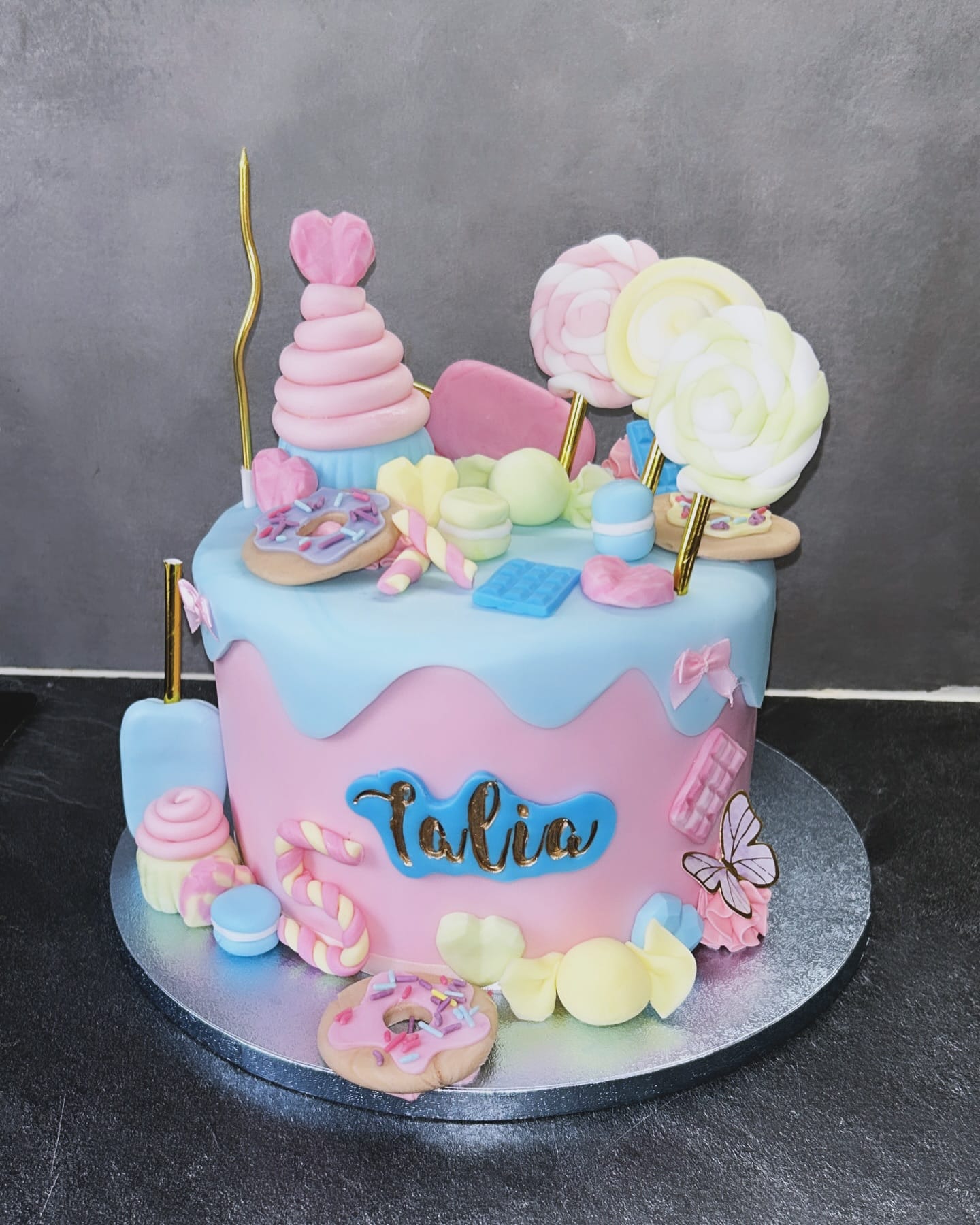 Gâteau d'anniversaire rose et bleu ciel, style "dripping" et surchargé de décorations de bonbons, macarons, beignets et sucettes. Inscription "Talia".