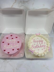 Deux petits gâteaux d'anniversaire ronds dans des boîtes blanches : un rose avec des cœurs rouges et écriture blanche "Happy Birthday", l'autre crème avec décoration festons roses et écriture rose.