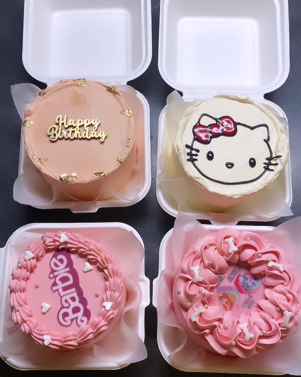 Quatre mini-gâteaux dans des boîtes blanches : un pêche avec "Happy Birthday", un Hello Kitty, un rose "Barbie" et un rose décoré avec des os de chiot.