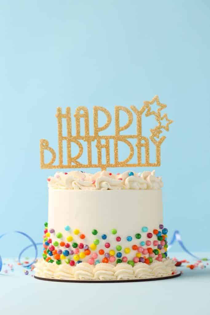 Gâteau d'anniversaire blanc avec décorations colorées et topper doré scintillant "HAPPY BIRTHDAY" sur fond bleu clair.