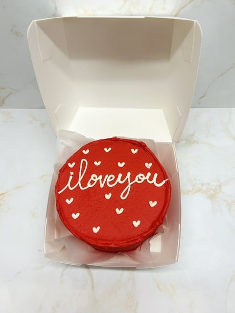 Mini gâteau rond rouge intense "i love you" en glaçage blanc et petits cœurs, présenté dans sa boîte ouverte sur marbre.