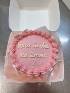 Gâteau rose pailleté dans une boîte blanche, glaçage blanc « Veux tu être ma témoin ? », rubans argentés et volutes roses. Une demande spéciale.