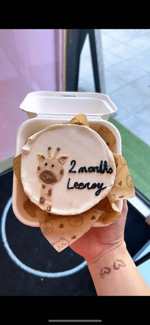 Mignon gâteau blanc "2 months Leeroy" avec girafe, dans emballage brun à motifs ours, tenu à la main pour célébrer 2 mois.