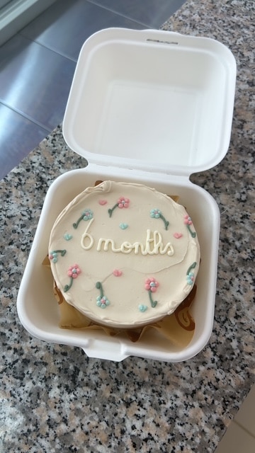 Bento cake blanc "6 months" décoré de fleurs pastel et cœurs dans sa boîte ouverte, posé sur un comptoir en granit moucheté.