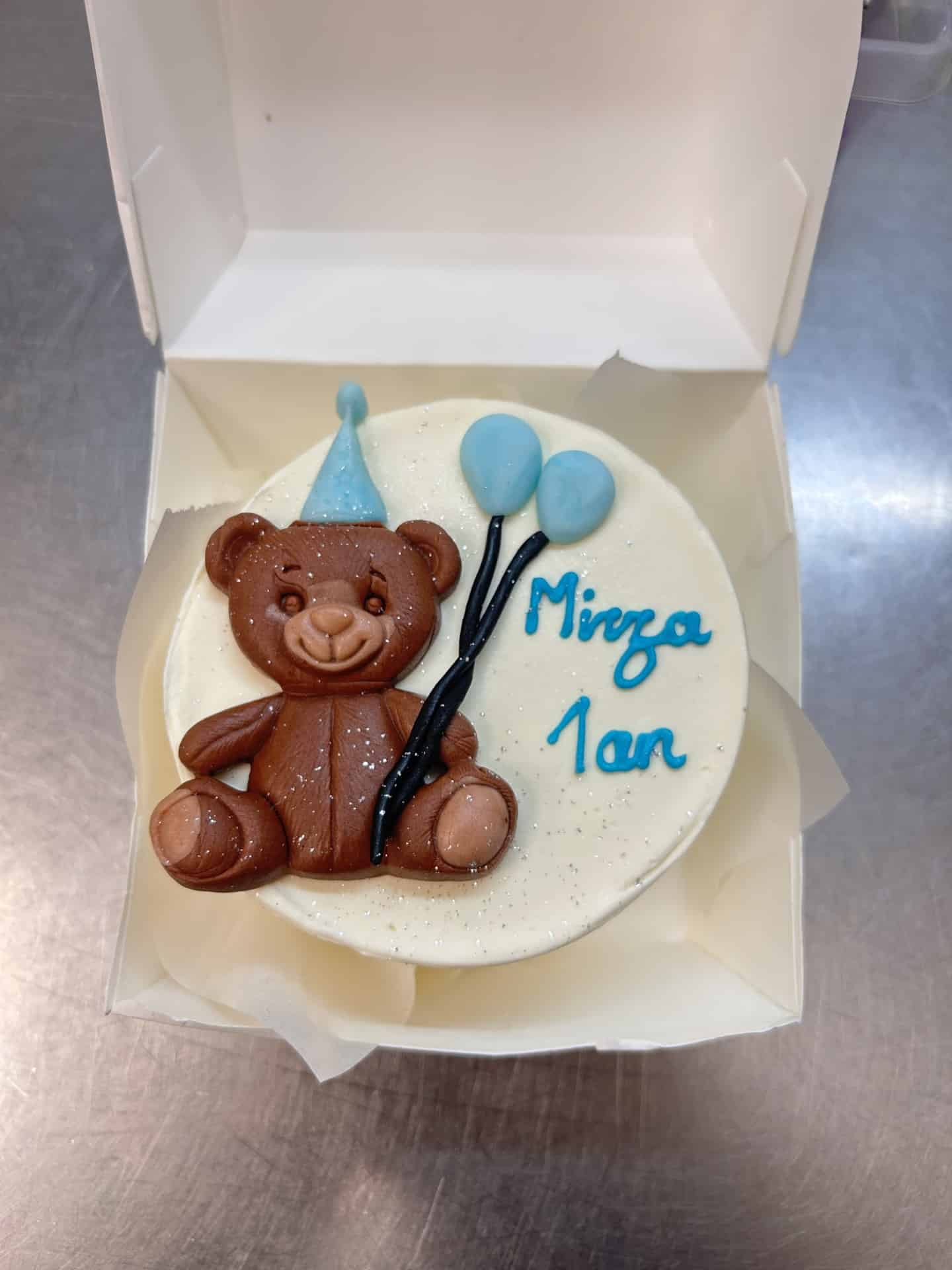Petit gâteau d'anniversaire blanc avec un ourson en chocolat, chapeau bleu et ballons. Le gâteau porte l'inscription "Mirza 1 an" et est saupoudré de paillettes, présenté dans une boîte ouverte.