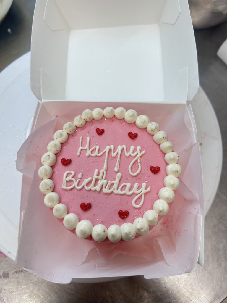Gâteau d'anniversaire rose vif avec glaçage blanc 'Happy Birthday', cœurs rouges et bordure de perles blanches, dans sa boîte.
