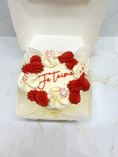 Gâteau blanc rond "Je t'aime" en glaçage rouge, décoré de volutes de crème blanche, de gouttes rouges et de cœurs roses, dans une boîte.