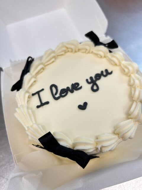Gâteau blanc rond "I love you" avec cœur noir et nœuds en ruban. Décoration élégante et romantique dans sa boîte.
