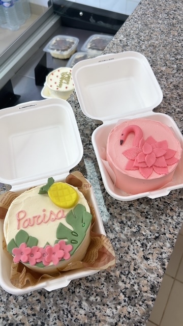 Trois petits gâteaux individuels, dont un rose flamant et un blanc avec "Parisa", ananas et fleurs, dans des boîtes à emporter.