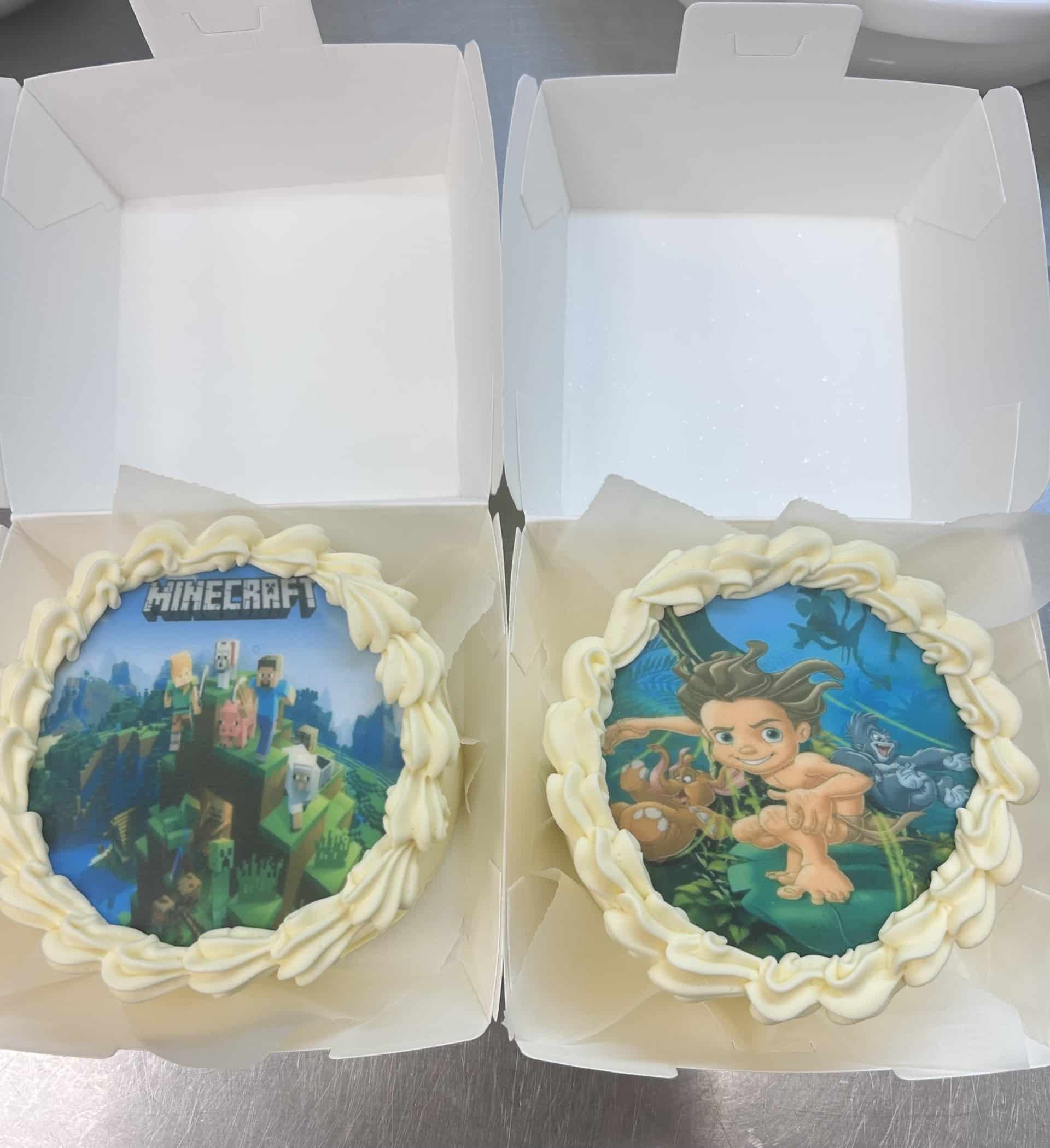 Deux gâteaux ronds individuels dans des boîtes blanches. L'un représente MINECRAFT avec son logo et personnages, l'autre Tarzan enfant dans la jungle avec des animaux.