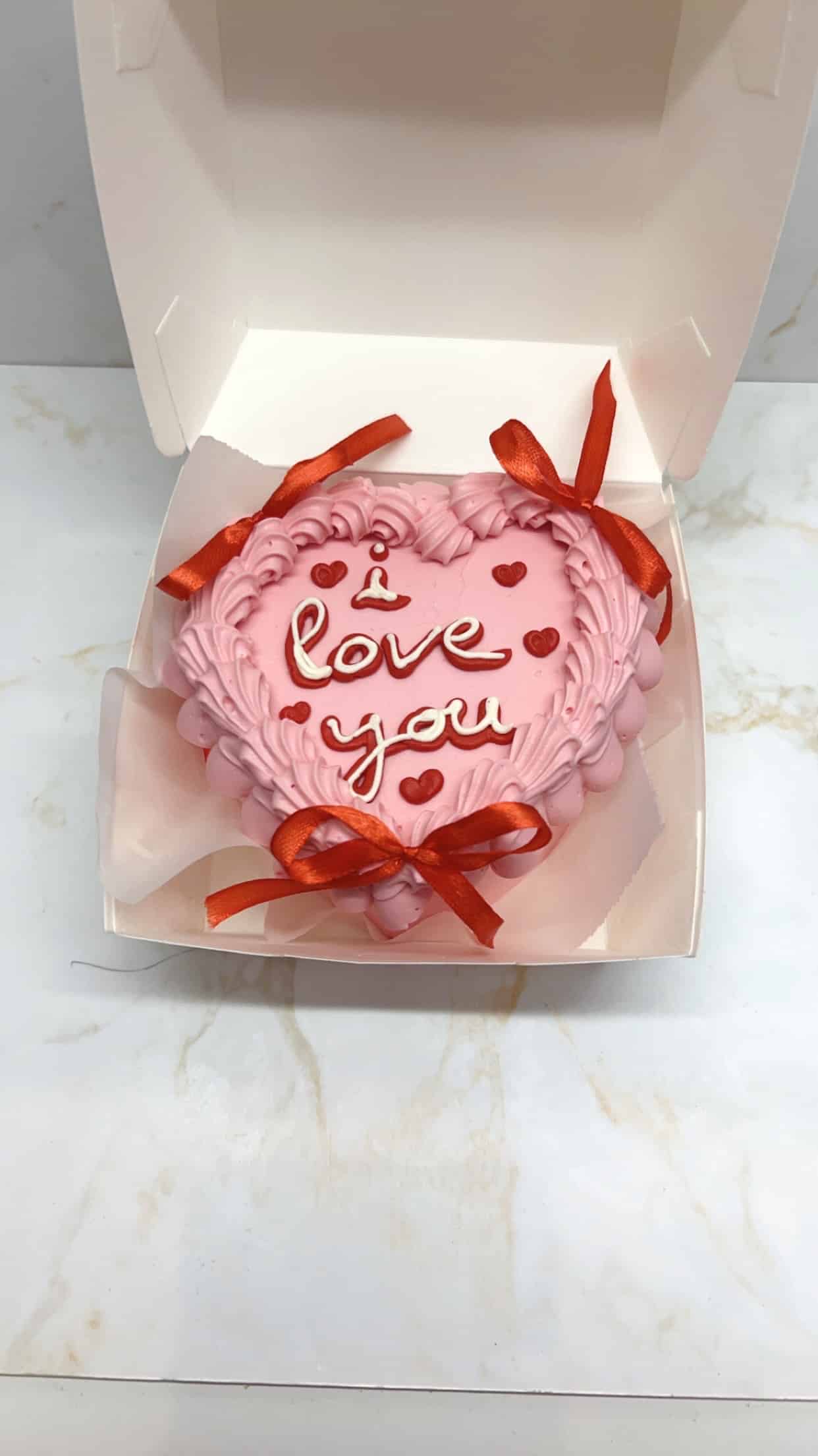 Gâteau cœur rose pâle "i love you" avec glaçage texturé, cœurs rouges et rubans. Présenté dans une boîte cadeau blanche sur marbre.