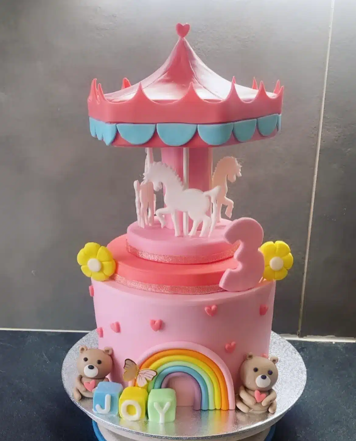 Gâteau d'anniversaire à trois étages sur le thème d'un carrousel rose avec chevaux blancs, un grand chiffre 3, des oursons, un arc-en-ciel et les lettres 'JOY'.
