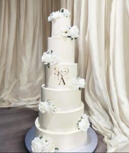 Gâteau de mariage blanc à cinq étages, décoré de roses blanches et d'un monogramme doré 'S & A', devant un fond de rideaux couleur ivoire.