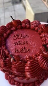 Gâteau rouge vif pailleté décoré de cerises et de glaçage texturé, avec l'inscription 'Older wiser hotter'.
