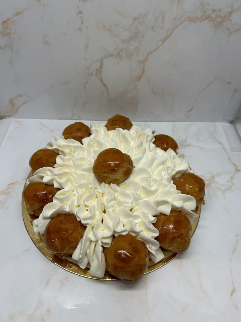 Gâteau Choux Crème Fouettée : Pâtisserie Exquise au Caramel Pâtisserie ronde de type Saint-Honoré recouverte de crème fouettée et de choux à la crème glacés au caramel, sur un plateau doré.