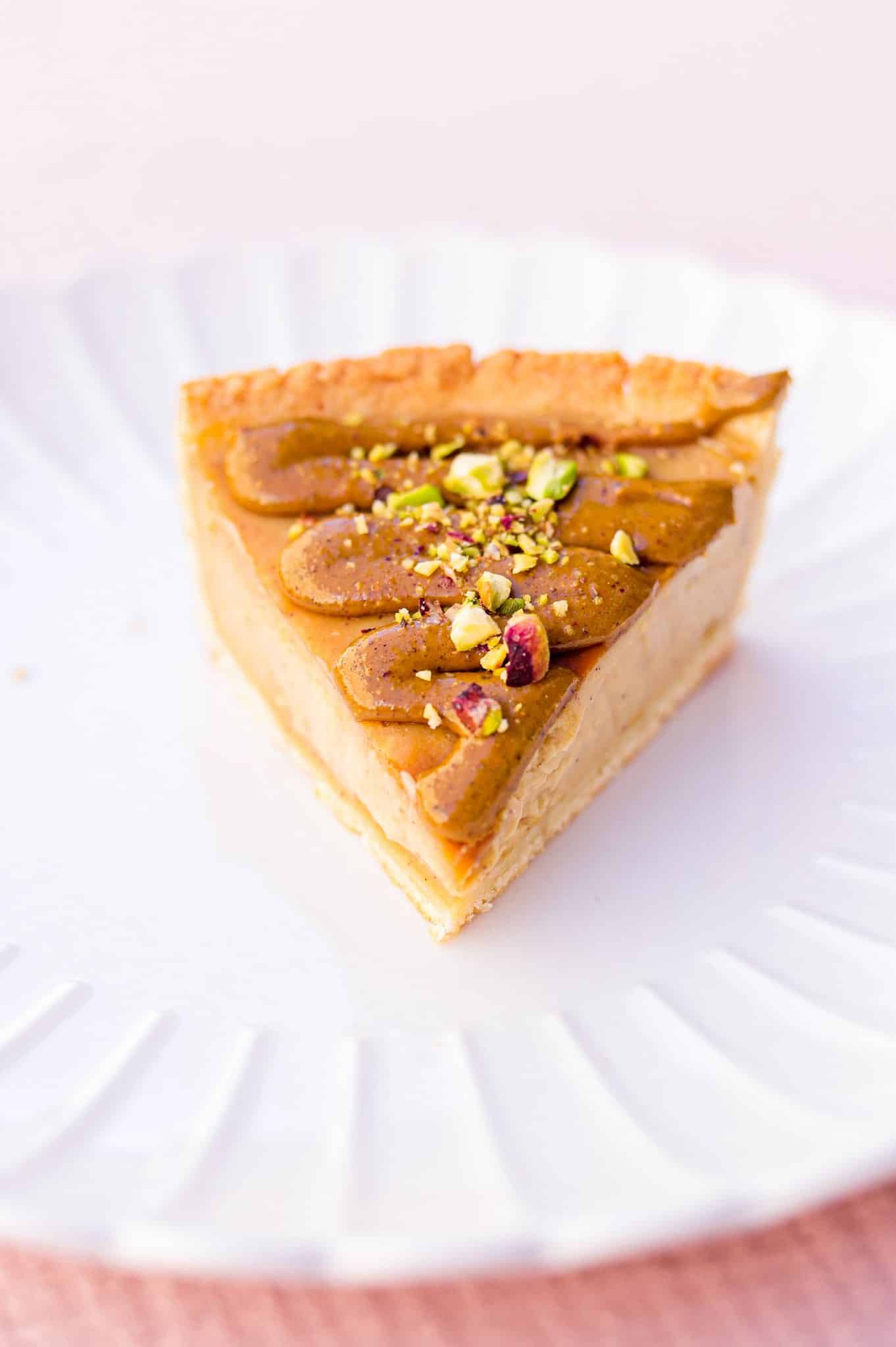 Part de tarte au caramel et pistaches sur une assiette blanche cannelée, fond rose poudré.