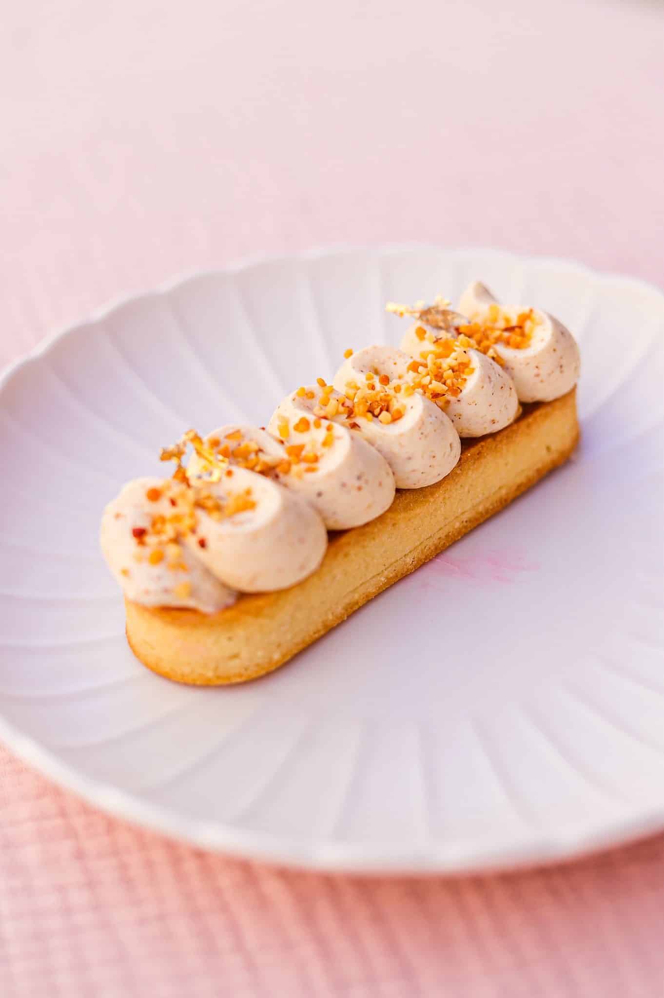 Tartelette allongée noisette et crème, éclats de noisettes et feuille d'or sur assiette blanche design.